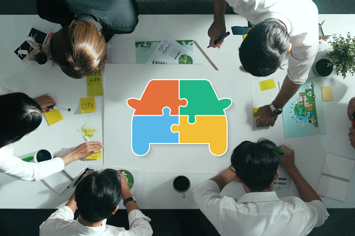 Personen sitzen um einen Tisch und arbeiten gemeinsamt. In der Mitte des Tisches ist eine bunte Grafik in Form eines Autos, die wie ein Puzzle zusammengesetzt ist.
