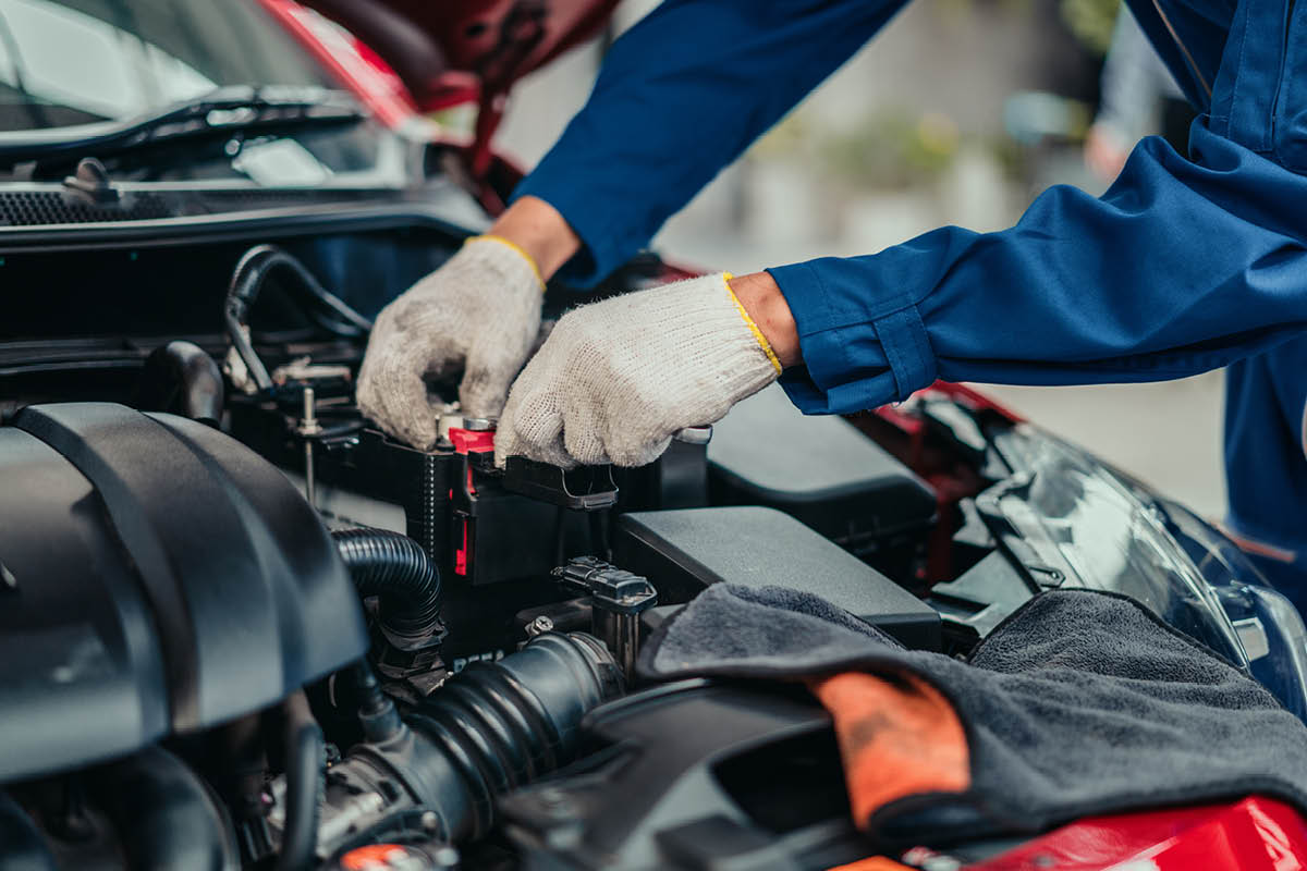 Eine Person in blauer Arbeitskleidung und Handschuhen arbeitet im Motorraum eines Autos an der Batterie. Verschiedene Motorkomponenten und Werkzeuge sind sichtbar.